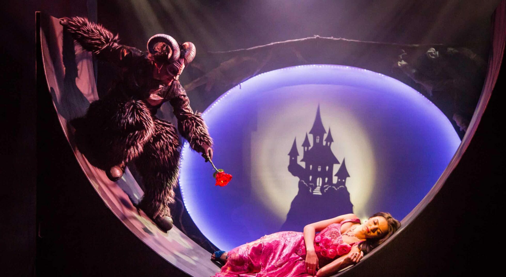 Man in Beast costume holds rose near sleeping woman.